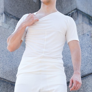 Puede incluir: Un hombre con una camiseta blanca y pantalones blancos se para frente a una pared de piedra gris.