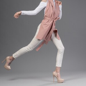 May include: A woman wearing a white shirt and a pink scarf tied at the waist, white pants, and nude heels. Her hair is pulled back in a ponytail and she is walking with her leg extended.