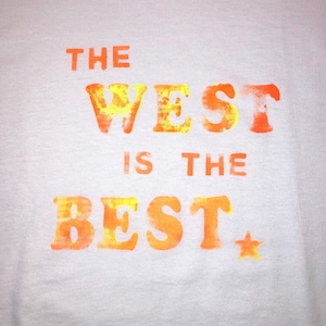 May include: White t-shirt with the text "THE WEST IS THE BEST" in orange and yellow. A small orange star is next to the word "BEST". The text has a textured, watercolor-like appearance.