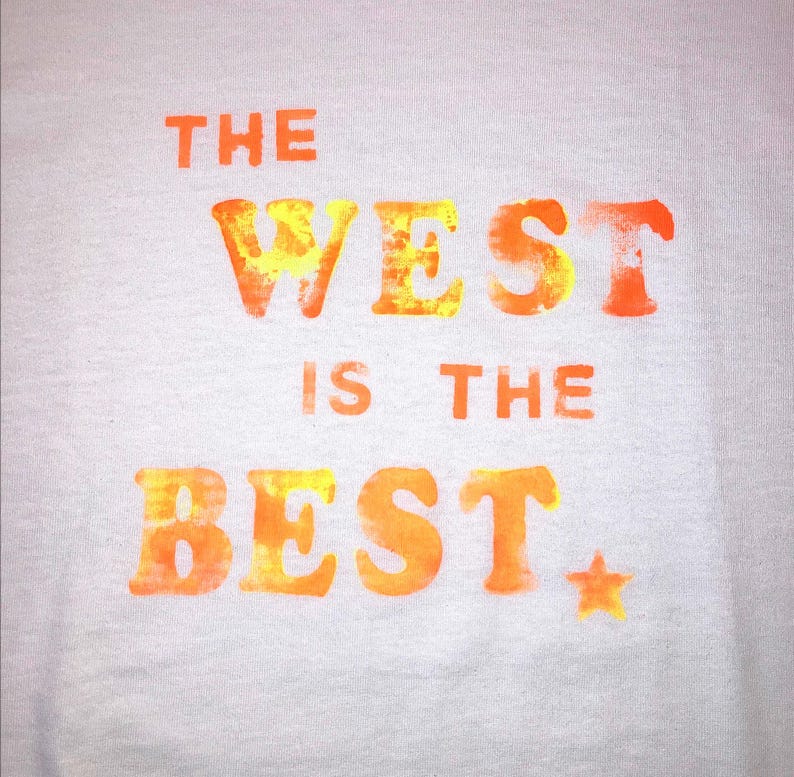 May include: White t-shirt with the text "THE WEST IS THE BEST" in orange and yellow. A small orange star is next to the word "BEST". The text has a textured, watercolor-like appearance.