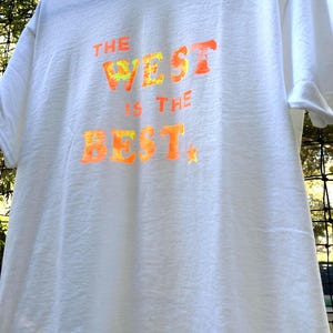 May include: White t-shirt with the text "THE WEST IS THE BEST" in orange and yellow tie-dye style lettering. A small yellow star is below the word "BEST". The shirt is hanging on a black hanger.