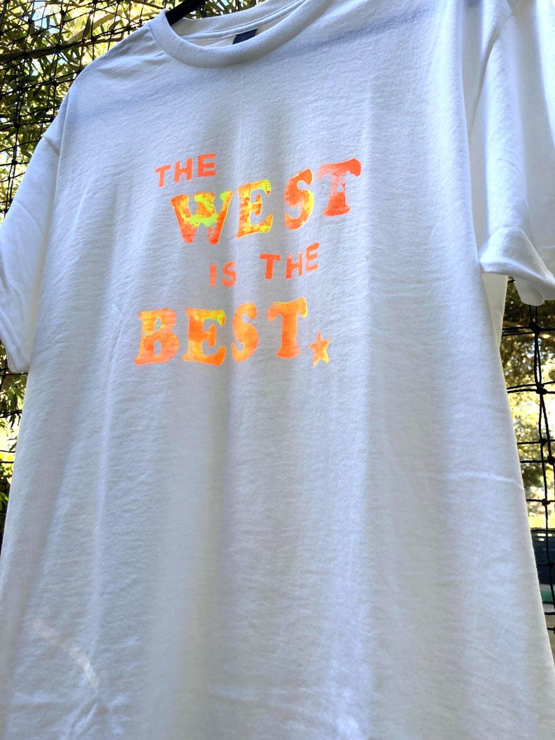 May include: White t-shirt with the text "THE WEST IS THE BEST" in orange and yellow tie-dye style lettering. A small yellow star is below the word "BEST". The shirt is hanging on a black hanger.