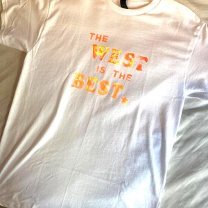 May include: White t-shirt with the text "THE WEST IS THE BEST" in orange and yellow lettering. The shirt has a crew neck and short sleeves. The text has a star at the end.