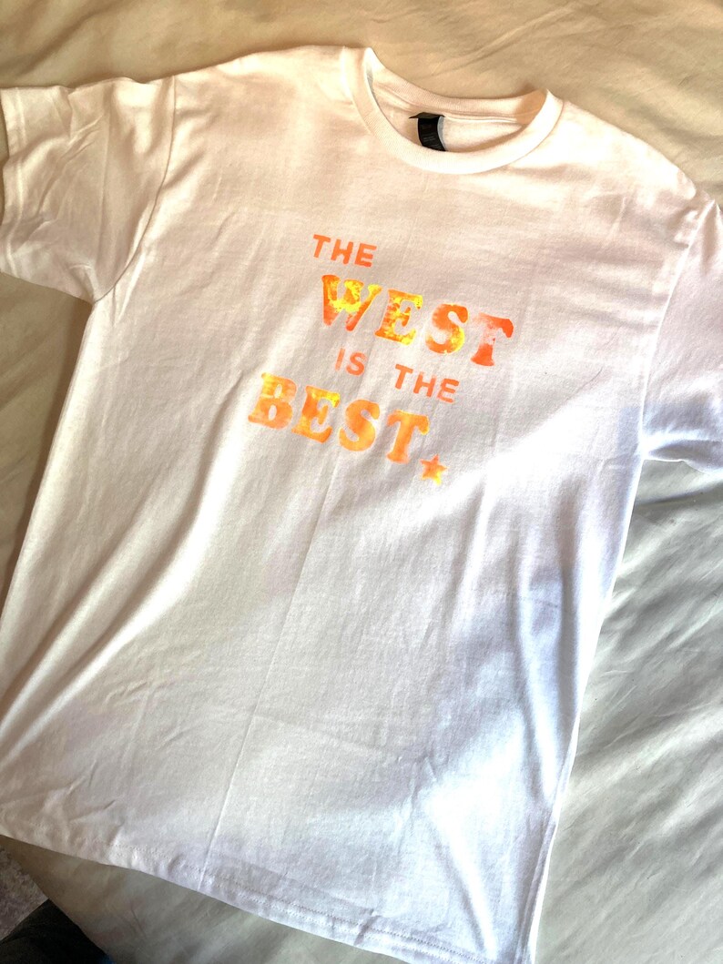 May include: White t-shirt with the text "THE WEST IS THE BEST" in orange and yellow lettering. The shirt has a crew neck and short sleeves. The text has a star at the end.