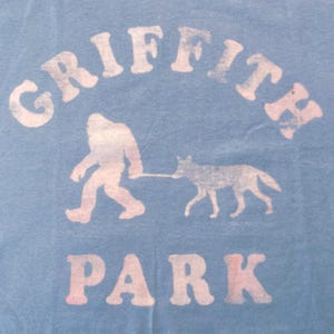 May include: Pale blue t-shirt with a graphic of Bigfoot walking a coyote on a lead. The text "GRIFFITH PARK" is arched above and below the figures in a faded white font.