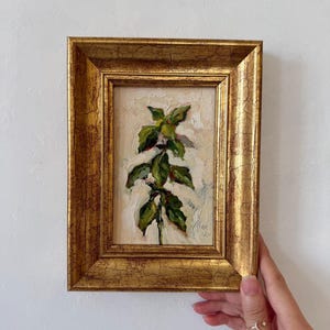 May include: An oil painting of a green basil plant with thick, textured brushstrokes on a cream-colored background, housed in a rectangular, ornate gold frame with a distressed, crackled finish and a signature in the bottom right corner.