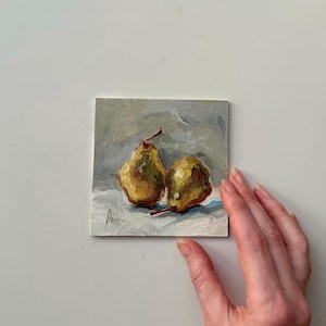 Op de afbeelding: Een klein schilderij van twee gele peren op een witte achtergrond. De peren zijn geschilderd in een realistische stijl met zichtbare penseelstreken. Het schilderij is onderaan links gesigneerd met "Ann".