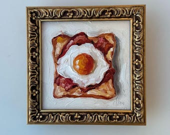 May include: An oil painting of a fried egg on toast with bacon, rendered in thick impasto brushstrokes. The artwork is encased in an ornate, antique-style gold frame with intricate scrollwork patterns along the edges and a white background.