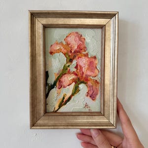 Könnte beinhalten: Ein Ölgemälde von zwei rosafarbenen Irisblüten mit dicken, strukturierten Pinselstrichen vor einem blassgrünen Hintergrund. Das Kunstwerk ist in einem metallisch goldenen Rahmen mit abgestuftem Profil gefasst und trägt eine kleine rote Signatur.