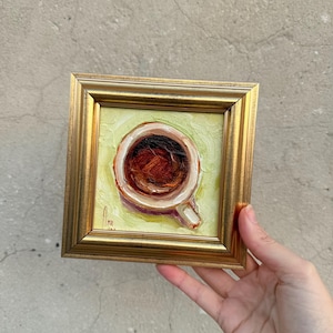 Op de afbeelding: Een klein vierkant olieverfschilderij in een goudkleurige lijst met een bovenaanzicht van een koffiekopje gevuld met donkerbruine vloeistof. De achtergrond van het schilderij is getextureerd met lichtgele en groene penseelstreken en een handtekening.