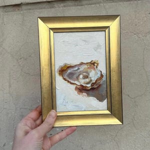 May include: A small painting of an oyster shell with a pearl inside. The shell is painted in shades of brown and white, and the pearl is a creamy white. The painting is framed in a gold frame.