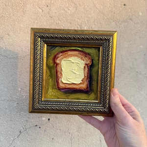 May include: A small square oil painting featuring a slice of toasted bread topped with a thick layer of yellow butter. The artwork is encased in an ornate, textured gold-toned frame with a rope-like border and a dark, olive-green background.