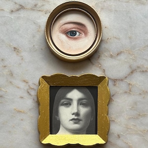 May include: A circular gold frame containing a painting of a single human eye, positioned above a square gold frame with scalloped edges containing a black and white portrait of a person with long hair, both displayed as distinct wall art pieces.
