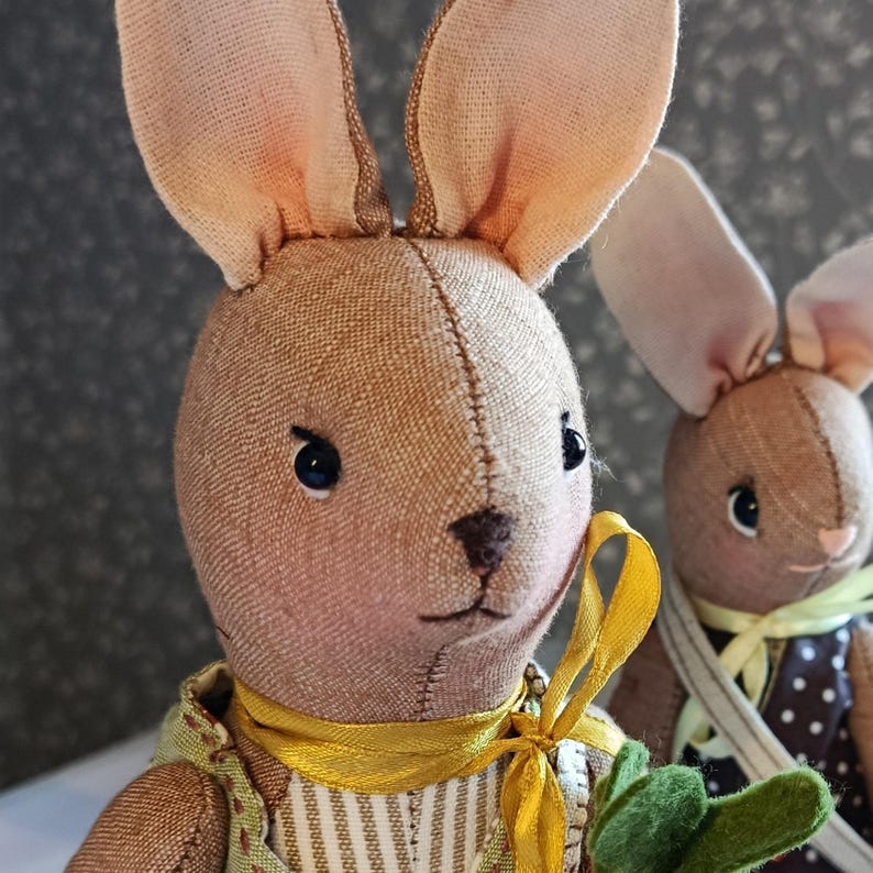 May include: Close-up of two handmade fabric bunny dolls. The bunnies have tan-colored bodies, long ears with pink accents, and black button eyes. One bunny wears a yellow ribbon and a green and yellow vest. The other bunny wears a brown and white polka dot dress.