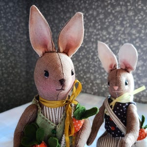May include: Two handmade fabric bunny dolls. The bunnies have brown bodies, pink-lined ears, and stitched facial features. One bunny wears a green vest with a yellow ribbon and holds carrot-shaped toys. The other bunny wears a polka-dot vest and a yellow scarf.