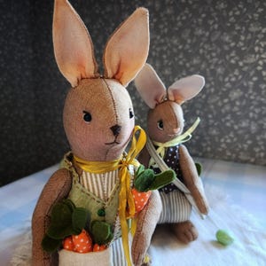 May include: Two handmade fabric bunny dolls. The larger bunny wears a green vest with a striped shirt and holds a carrot pouch. The smaller bunny wears a polka dot vest and striped pants. Both have long ears and are brown.