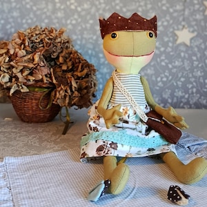 Könnte beinhalten: Eine handgefertigte Froschpuppe mit einem braunen Cord-Kronen, die ein geblümtes Kleid mit blauem Besatz und ein gestreiftes Hemd trägt. Die Puppe hat eine braune Umhängetasche und sitzt auf einem karierten Stoff. Ein Korb mit getrockneten Blumen befindet sich im Hintergrund.