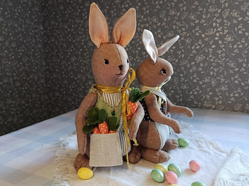 May include: Two handmade Easter bunny dolls with long ears, wearing vests and carrying a bag of carrots. The bunnies are brown and tan, with yellow and green accents. Colorful Easter eggs are scattered around the bunnies on a white cloth.