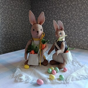 May include: Two handmade Easter bunny dolls with long ears, wearing aprons and holding carrots. One bunny has a striped apron and the other has a polka dot apron. Pastel colored Easter eggs are scattered around the bunnies.