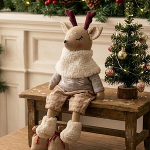 May include: A plush deer doll with dark red antlers, wearing a white sherpa fleece collar, a grey long-sleeved shirt, and plaid trousers. The doll has embroidered closed eyes, pink cheeks, and cream-colored fuzzy boots, sitting on a wooden bench.