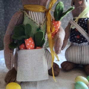 May include: Two handmade Easter bunny dolls. One bunny wears a green vest and carries a bag of felt carrots with white polka dots. The other bunny wears a black and white polka dot vest. Both bunnies have brown fur and are decorated with yellow ribbons.