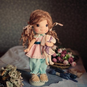 May include: A handmade doll with brown curly hair in pigtails, wearing a pink dress, blue pants, and tan shoes. The doll is holding a stuffed bunny. The doll is standing on a white base.