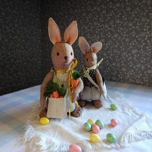May include: Two brown fabric bunny dolls with long ears, dressed in aprons and vests, holding carrots. Pastel Easter eggs are scattered on a white cloth. The bunnies are on a table with a blue and white checkered tablecloth.