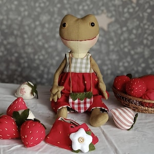 May include: A stuffed frog doll with a tan body wearing a red dress with a white gingham bodice and a matching strawberry-shaped crossbody bag. The doll is surrounded by several fabric strawberries and a red accessory with a white flower.