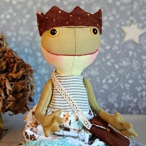 Könnte beinhalten: Handgemachte Froschpuppe mit brauner Krone, gestreiftem Hemd und Blumenkleid. Die Puppe hat eine kleine braune Handtasche und einen weißen Spitzenträger. Der Frosch hat große gelbe Augen und ein gesticktes Lächeln mit rosigen Wangen.
