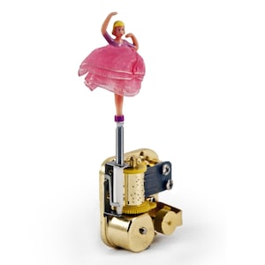 May include: A gold-colored music box with a ballerina figurine on top. The ballerina is wearing a pink dress and has blonde hair. The music box has a winding key and a small, round opening on the side.