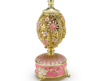 Op de afbeelding: Een goudkleurige, sierlijke eivormige muziekdoos met roze bloemaccenten. Het ei heeft een filigraan ontwerp en staat op een roze en gouden voet met decoratieve voetjes. De bovenkant van het ei is ook goudkleurig en heeft een decoratieve bekroning.