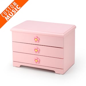 Puede incluir: Una caja de joyería de madera rosa con tres cajones. Cada cajón tiene una perilla con forma de flor con un centro amarillo.