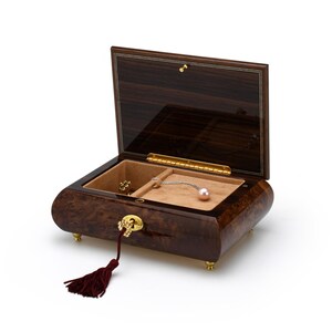 Puede incluir: Una caja de m&uacute;sica de madera marr&oacute;n oscuro con un borla roja. La tapa est&aacute; abierta, revelando un interior de terciopelo beige con un peque&ntilde;o compartimento y un collar de perlas.