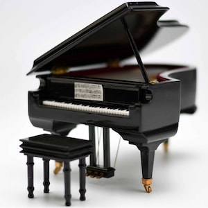 Pode incluir: Um piano de cauda em miniatura preto com uma partitura musical dentro e um banquinho de piano preto correspondente. O piano tem uma guarni&ccedil;&atilde;o dourada e o banquinho tem quatro pernas.