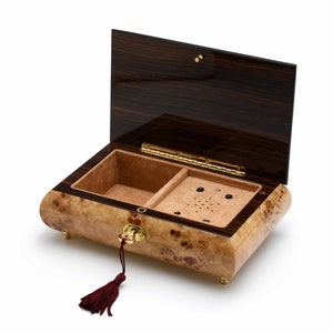May include: A wooden music box with a dark wood lid and a light wood base. The lid is open and the interior is lined with brown felt. The music box has a key and a tassel.