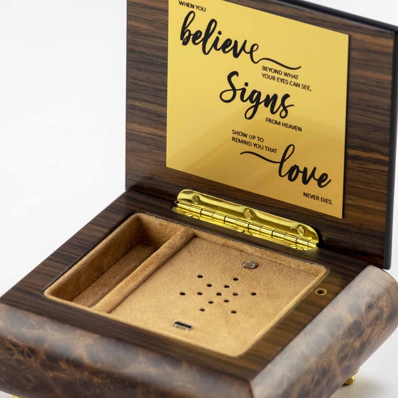 Puede incluir: Una caja de m&uacute;sica de madera con una placa dorada que dice "Cuando crees en las se&ntilde;ales del cielo, aparecen para recordarte que el amor nunca muere". La caja est&aacute; abierta y tiene un interior de terciopelo marr&oacute;n.