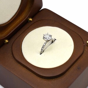 May include: A diamond engagement ring with a round brilliant cut center stone and a pave diamond band, displayed in a wooden ring box.