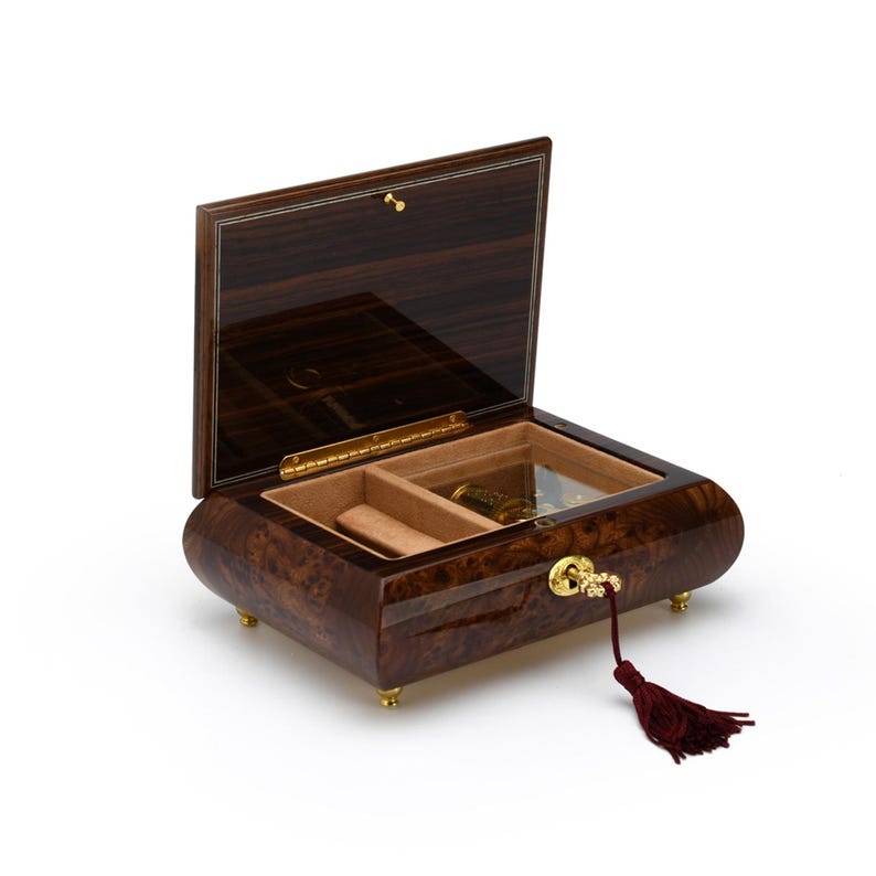 Puede incluir: Una caja de m&uacute;sica de madera con un acabado marr&oacute;n oscuro y una llave dorada. La caja est&aacute; abierta, revelando un peque&ntilde;o compartimento y un mecanismo de cuerda. La caja tiene un borla atada a la llave.