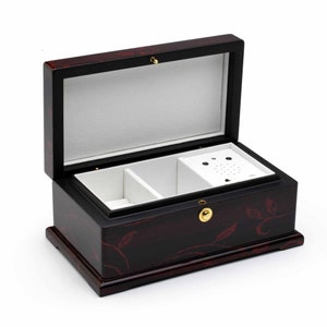 以下が含まれることがあります： 花柄の黒い木製のオルゴール。蓋が開いており、白いベルベットの内側に2つの仕切りと小さな音楽プレーヤーが見える。