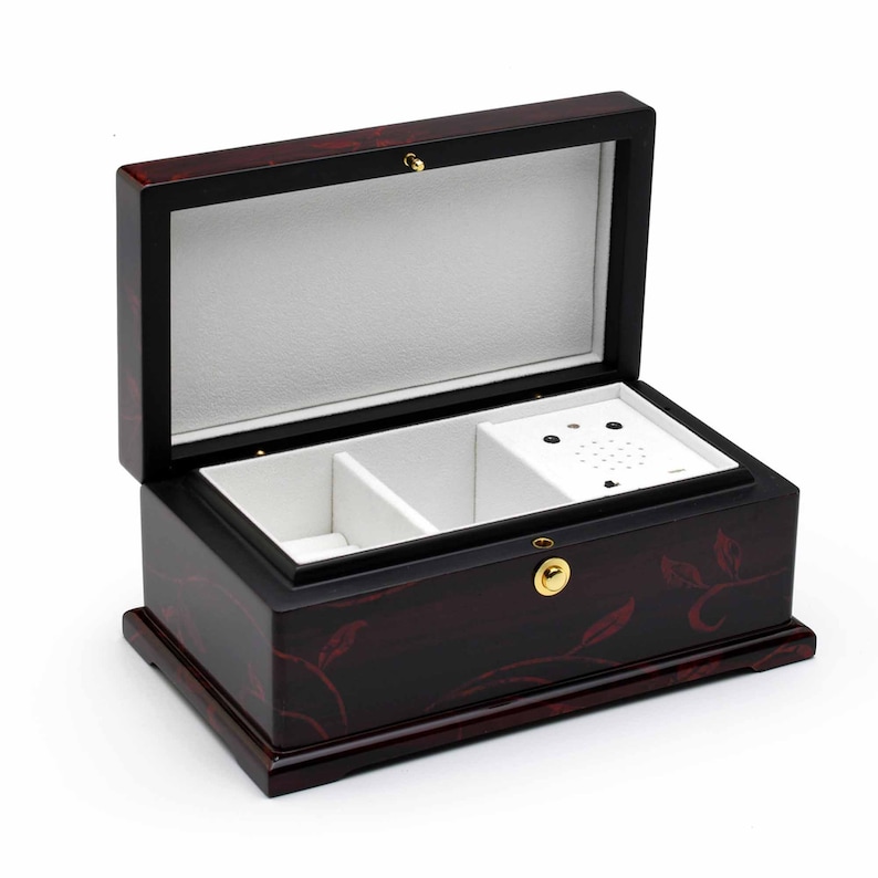 以下が含まれることがあります： 花柄の黒い木製のオルゴール。蓋が開いており、白いベルベットの内側に2つの仕切りと小さな音楽プレーヤーが見える。