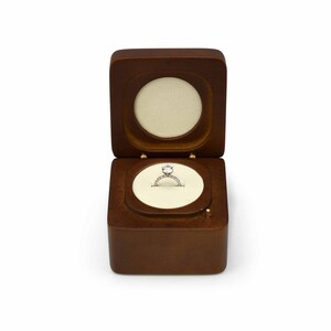 May include: A wooden ring box with a hinged lid, open to reveal a diamond engagement ring on a white velvet cushion.