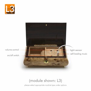 May include: A wooden music box with a brown lid and a light wood interior. The box has a volume control knob, an on/off switch, a light sensor, and a USB port for self-loading music. The text "(module shown: L3)" and "please select appropriate module type under options" is printed below the box.