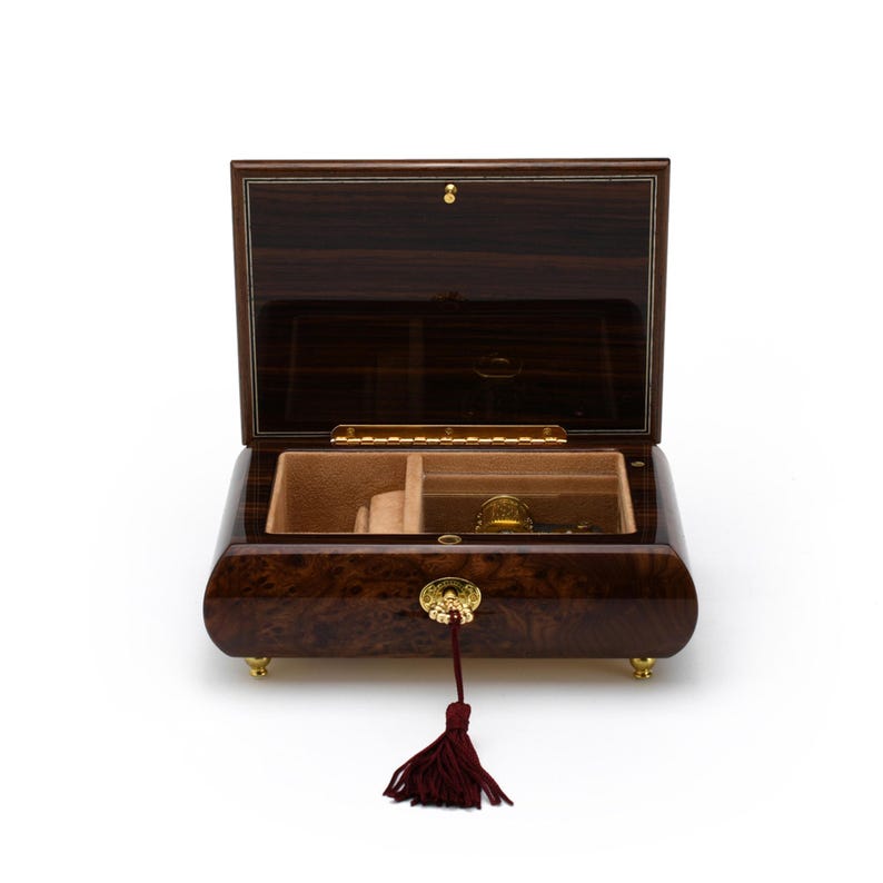 Puede incluir: Una caja de m&uacute;sica de madera oscura con un borla de color burdeos. La tapa est&aacute; abierta, revelando un interior beige con un compartimento y un mecanismo de cuerda.