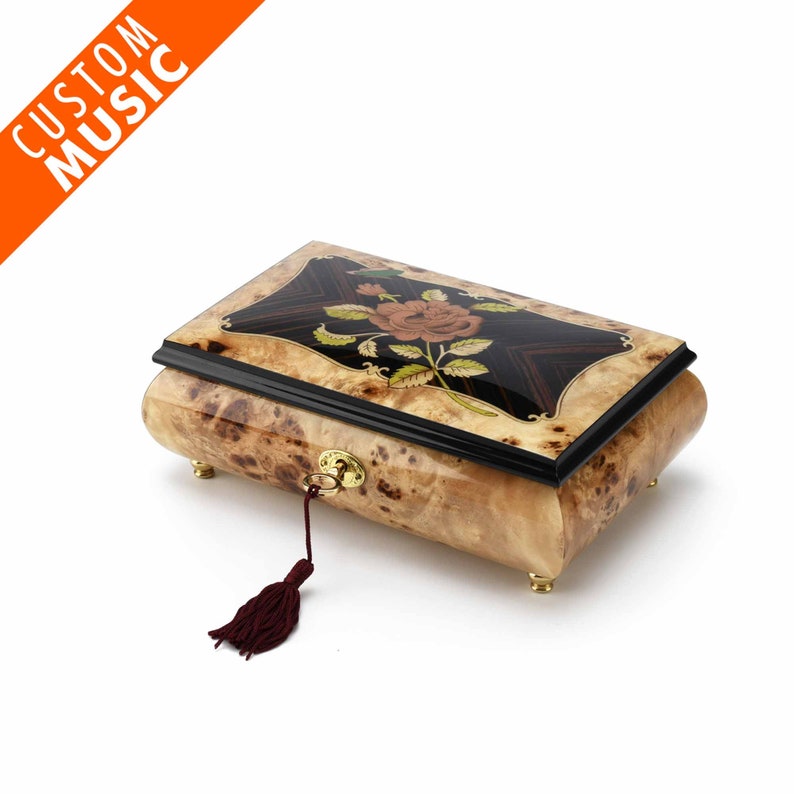 May include: A wooden music box with a floral inlay design. The box is closed and has a keyhole with a tassel. The box is made of light wood with dark wood accents.