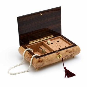May include: A wooden music box with a burl wood finish and a dark wood lid. The box is open and contains a pearl necklace, a ring, and a pair of earrings. The box has a keyhole and a tassel.
