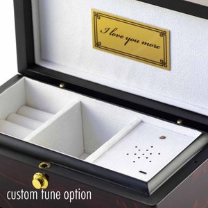 以下が含まれることがあります： 白いベルベットの内装が施された黒い木製のジュエリーボックス。ボックスにはリング用の小さなコンパートメントと、音楽を再生するためのスピーカー付きの大きなコンパートメントがあります。ボックスの蓋には、「もっと愛してる」と書かれた金色のプレートが付いています。