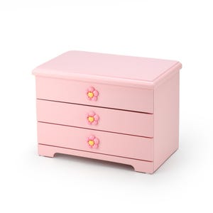 Puede incluir: Una cómoda de madera rosa con tres cajones y pomos con forma de flor. Los cajones están cerrados.