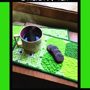 May include: A green quilted snack mat featuring a shamrock pattern, a mug of steaming dark liquid, and three dark round cookies. The text reads Shamrock Snack Mat, Tricia Lynn Maloney, Orphan Quilt Designs, and for Villa Rosa Designs.
