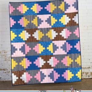 May include: A quilt featuring a repeating geometric block pattern in yellow, pink, blue, brown, and black and white gingham fabrics. The text THROWBACK appears at the top, and Running Doe QUILTS For Villa Rosa Designs with a doe graphic at the bottom.