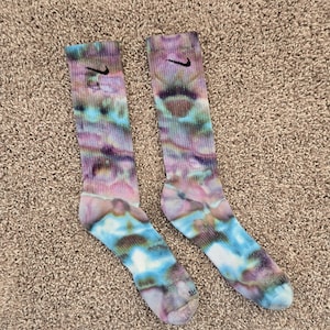 May include: A pair of crew-length socks featuring a tie-dye pattern in shades of blue, purple, and brown. Each sock has a ribbed cuff and a small black Nike swoosh logo embroidered near the top. The fabric displays a blended, marbled effect.